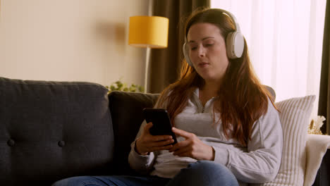 Woman-Wearing-Wireless-Headphones-Sitting-On-Sofa-At-Home-Streaming-Music-From-Mobile-Phone-2